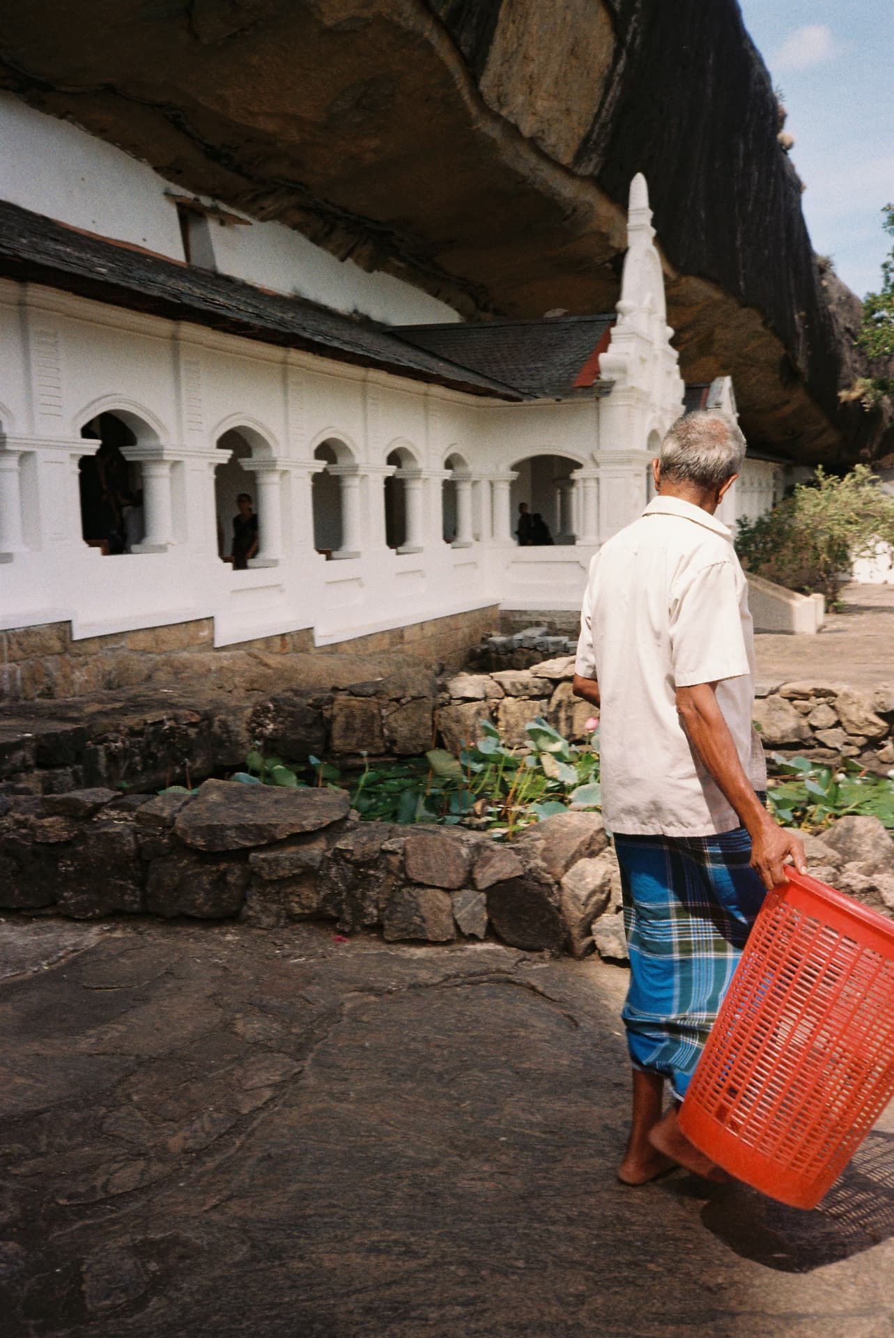 Man carrying a red basket past a whitewashed cave temple